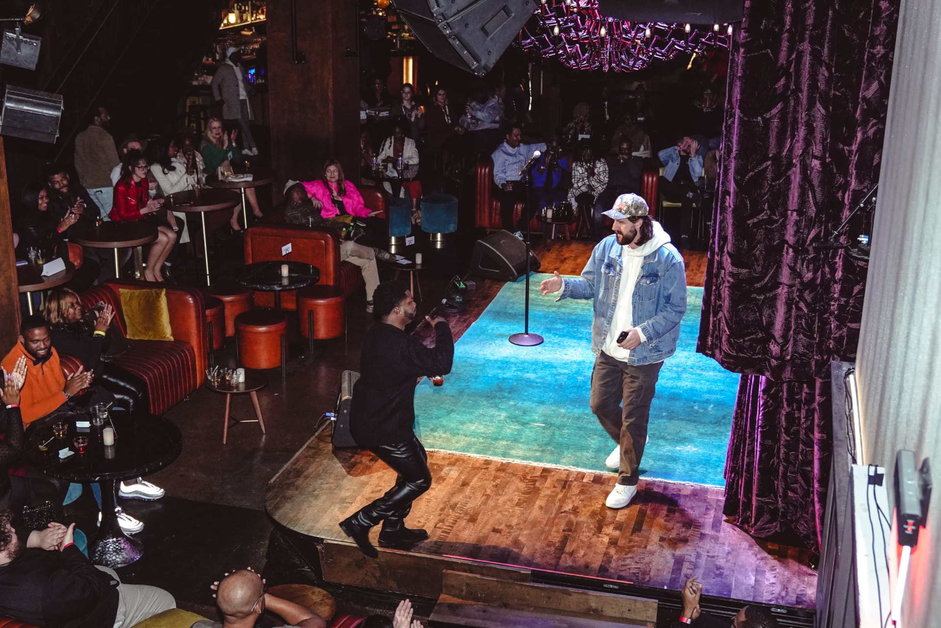 Two comedians greeting on a teal-carpeted stage seen from above