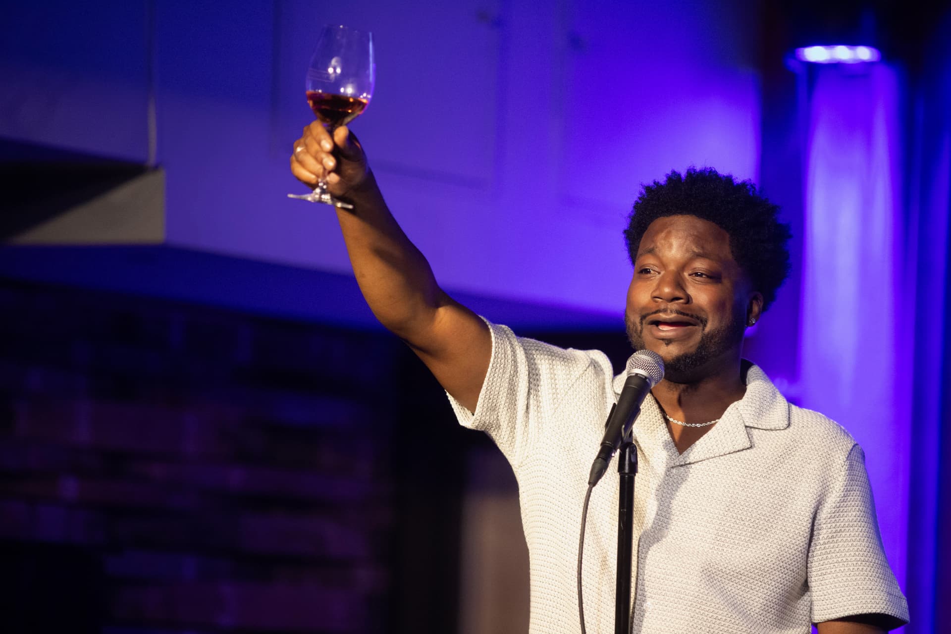 Comedian raising a glass of wine at the mic under purple stage lights